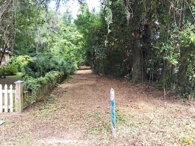 Cleared driveway with mulched ground, mature trees, and white picket fence entry